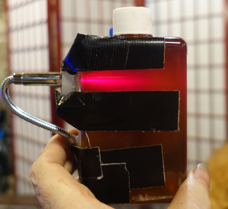 This photo shows my experimental Red PNSB Laser Device, with the laser turned on. Photo was shot in a normally-lighted room. The plastic centrifuge tube mounted on the back of the device contains an alchemically-created mixture of PNSB and certain minerals. The material in the tube provides a beneficial field effect/space effect that bathes the liquid PNSB culture in the bottle, along with the laser beam itself.